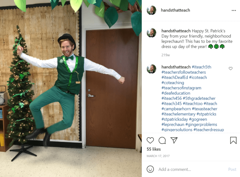 Teacher in a leprechaun outfit in a classroom jumping and clicking his heels in front of a Christmas tree with shamrock garland