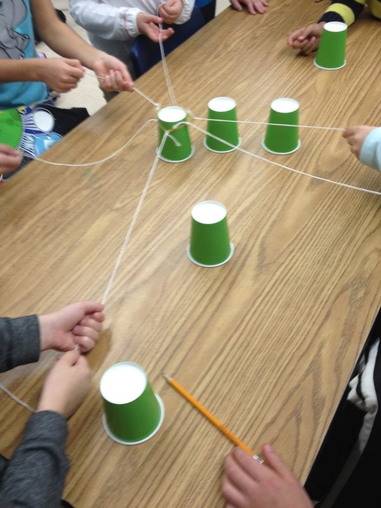 kids around a table playing a cup stacking game with paper cups and string