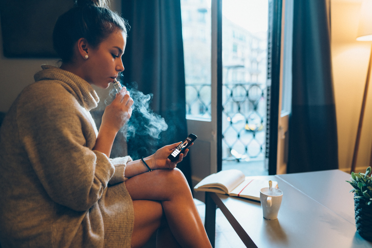 Teen girl vaping while sitting on a sofa and looking at a phone.