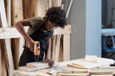 Waist up portrait of young black boy in carpentry workshop using power drill while building furniture, copy space