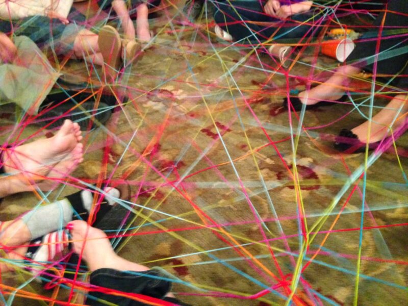 Students legs extended on a colorful rug in a circle, with strands of yarn connected in a web between them