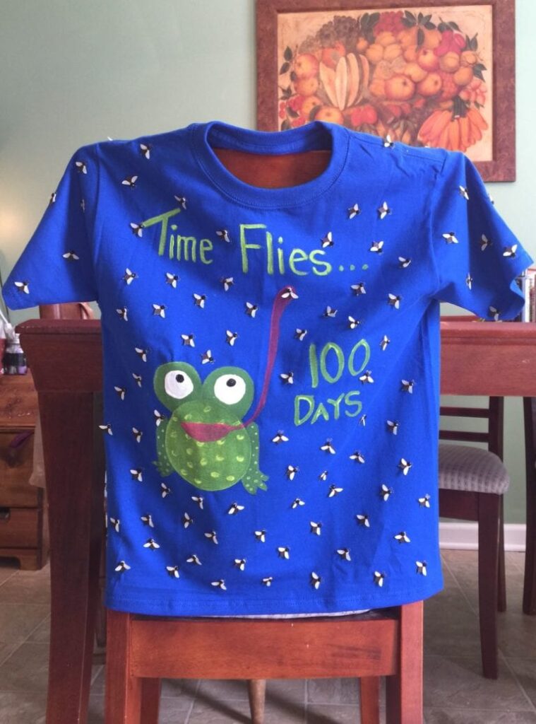 A blue homemade shirt says Time flies 100 days. It has a frog with a big tongue catching flies that are all over the shirt.