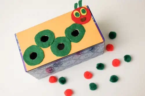 A tissue box has a caterpillar on the top of it. The body has holes in it. There are red and green pom poms to put inside the box. (very hungry caterpillar activities)