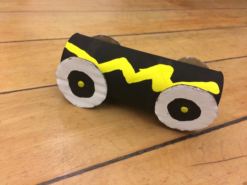 A toilet paper roll is painted black with a yellow lighting bolt. Cardboard wheels have been added.