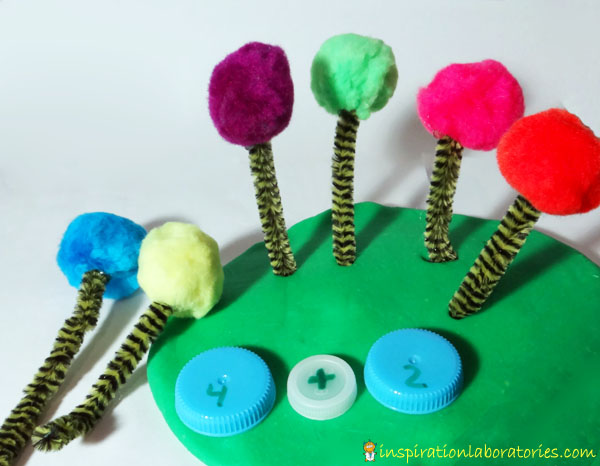 green play dough with trees made from pipe cleaners and pom poms stuck into it with 3 plastic bottle caps showing a math sentence