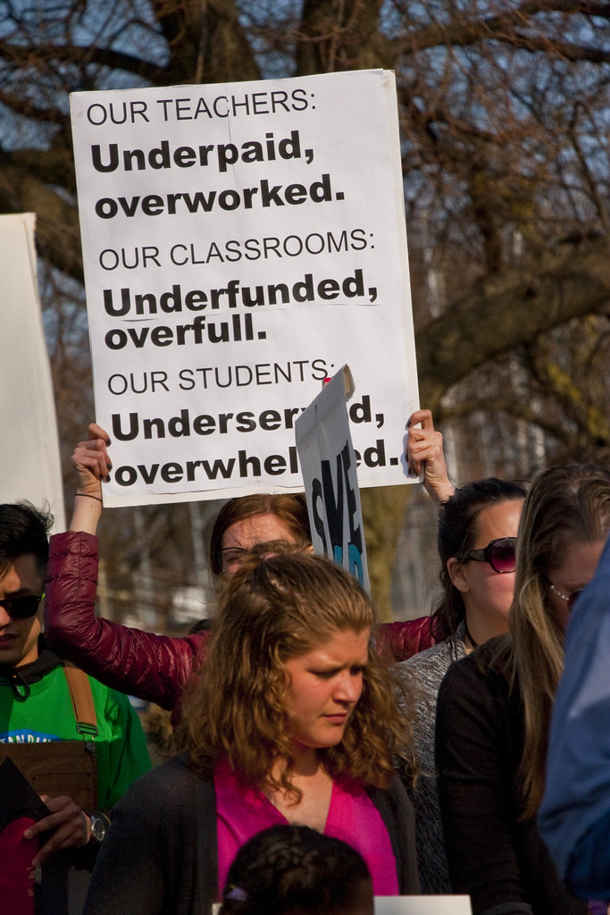 woman holding up a sign that says underfunded overworked 