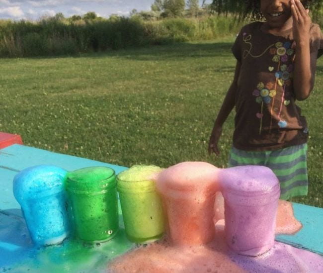 Girl looks on as rainbow colored foam erupts from glass jars (Volcano Science Experiments)