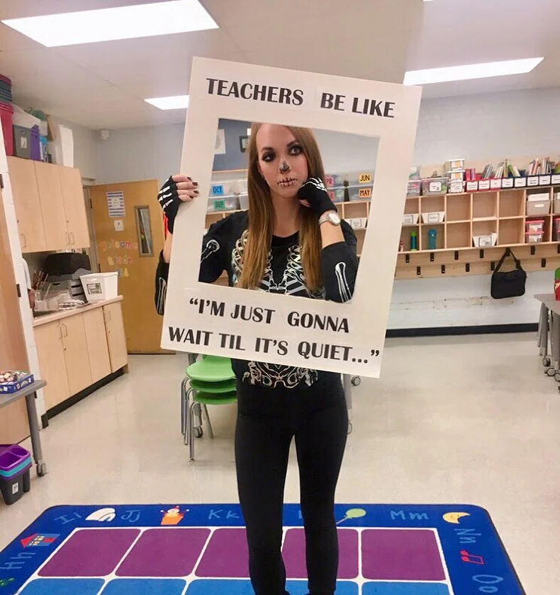 A woman is dressed as a skeleton and holding a frame around her face that says "teachers be like 'I'm just gonna wait til it's quiet.'"