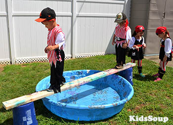 A long wooden board is across a kiddie pool and kids are walking across it dressed in pirate costumes.