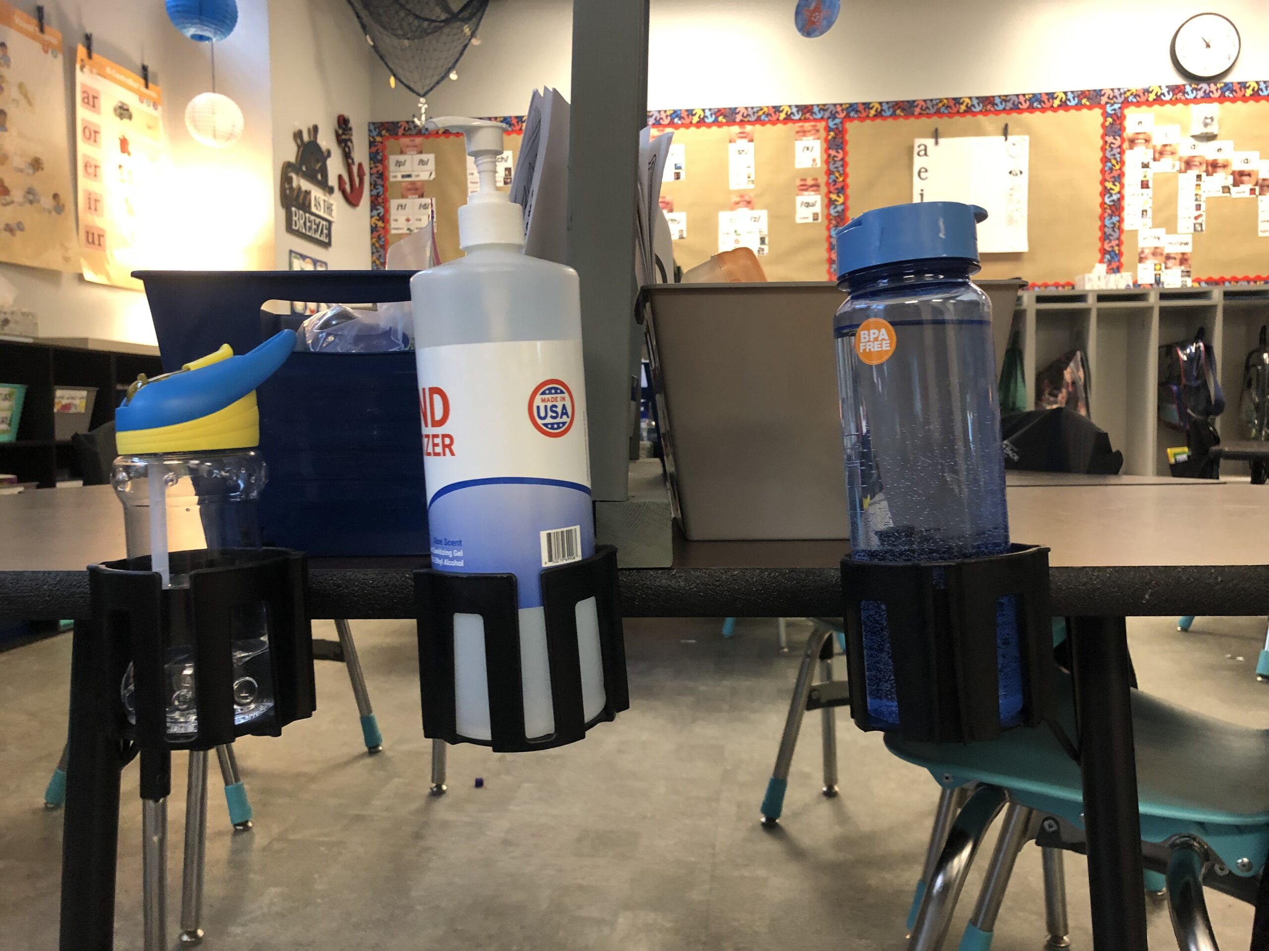 Water bottle holders for student desks include these cup holster bottle holders attached to aclassroom desk, holding water bottle and cleaning wipes 