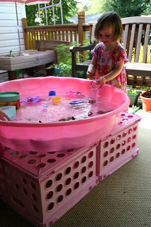 A pink kiddie pool is sitting on top of four large plastic crates and a child plays with water toys in it.