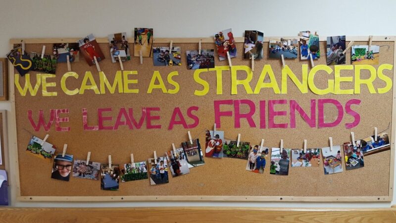 Text reads We Came as Strangers, We Leave as Friends. There are clotheslines with clothespins holding up pictures of students and teachers. 