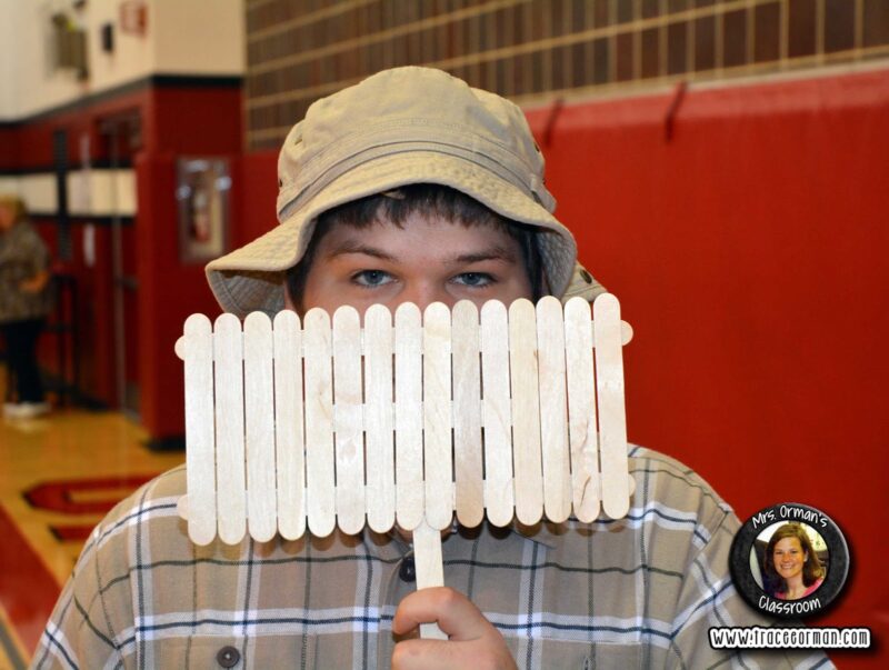 Teacher Halloween costumes can be from popular tv shows like this one. A boy wears a fishing hat and a plaid shirt and is holding a fence made from popsicle sticks in front of his face.