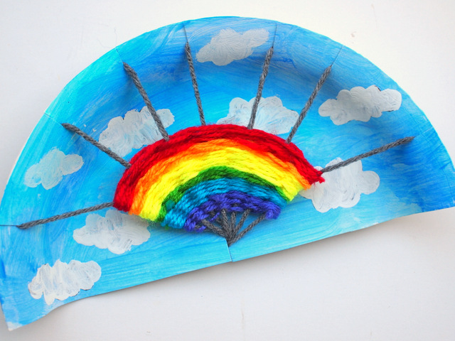 A paper plate is painted blue with white clouds. A rainbow has been woven in the center using different color yarn (spring crafts for kids)