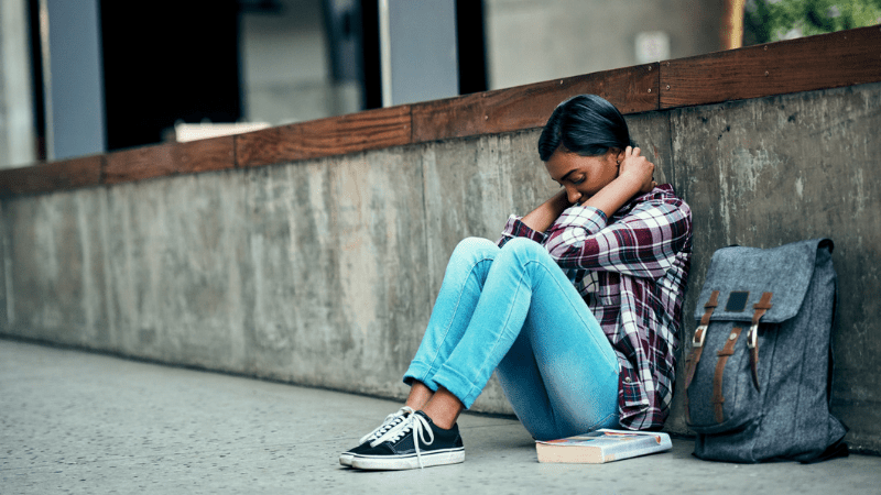 Adolescent girl who needs youth suicide prevention sitting outside school with head down